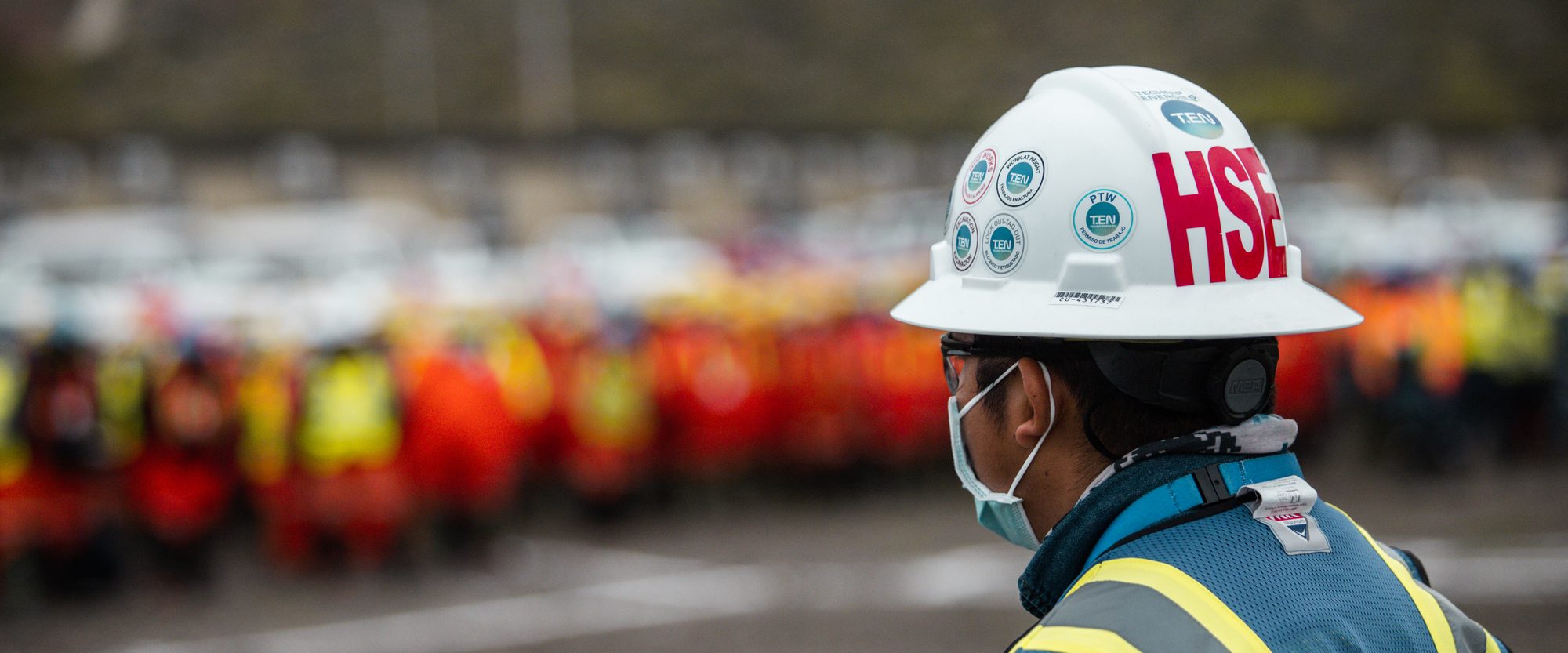 Pre-start briefing on an LNG construction site at first light: a supervisor in an HSE hard hat in the foreground, the incoming shift assembled behind him.
