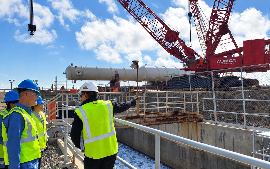 A group on a construction walkdown at Energ&iacute;a Costa Azul, one of them pointing out an item of plant, with a crawler crane and a pressure vessel behind them.